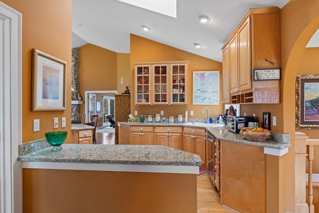 kitchen featuring a peninsula, a sink, light wood-style floors, vaulted ceiling with skylight, and glass insert cabinets