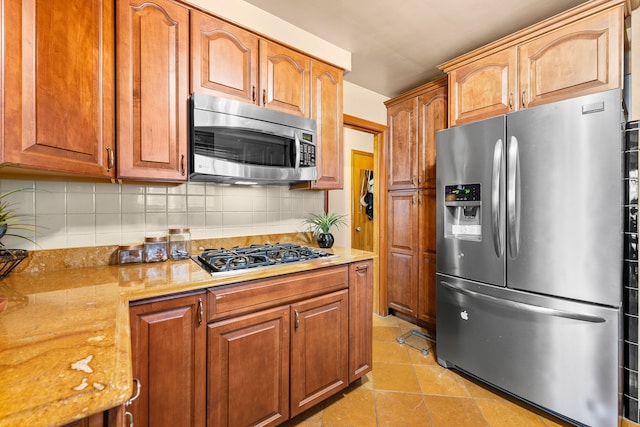 kitchen featuring stainless steel appliances, brown cabinetry, light tile patterned flooring, light stone countertops, and tasteful backsplash