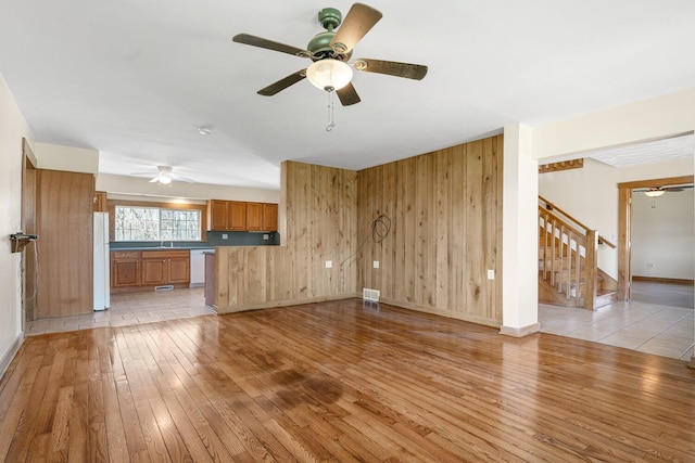 unfurnished living room with stairway, a ceiling fan, light wood-style flooring, wood walls, and visible vents