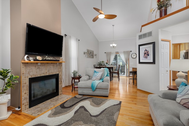 living room with high vaulted ceiling, a tiled fireplace, light wood-style flooring, a ceiling fan, and a chandelier