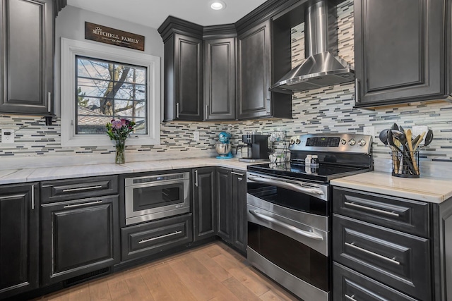 kitchen with tasteful backsplash, dark cabinets, range with two ovens, light wood-type flooring, and wall chimney exhaust hood