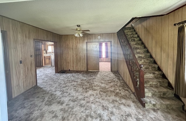 unfurnished living room with stairway, wooden walls, ceiling fan, and carpet flooring