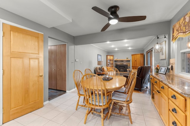 dining area with light tile patterned floors, ceiling fan, a stone fireplace, and baseboards