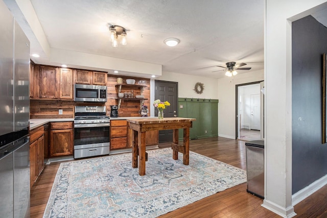 kitchen with dark wood finished floors, brown cabinetry, stainless steel appliances, baseboards, and a ceiling fan