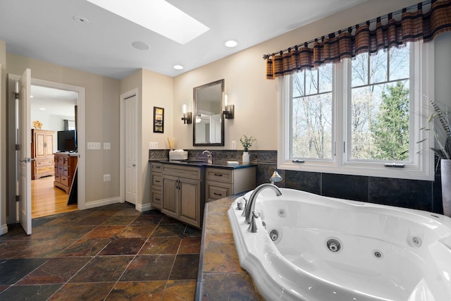 bathroom with stone tile flooring, vanity, a whirlpool tub, baseboards, and a skylight