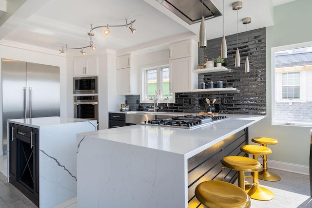 kitchen with decorative backsplash, open shelves, light stone countertops, built in appliances, and a breakfast bar