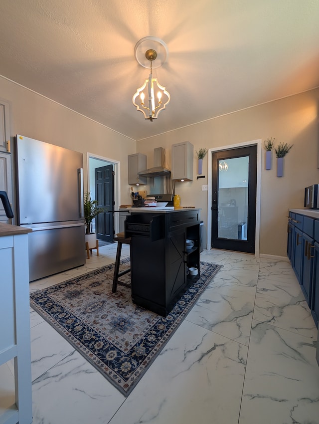 kitchen featuring a notable chandelier, marble finish floor, a kitchen island, wall chimney range hood, and freestanding refrigerator