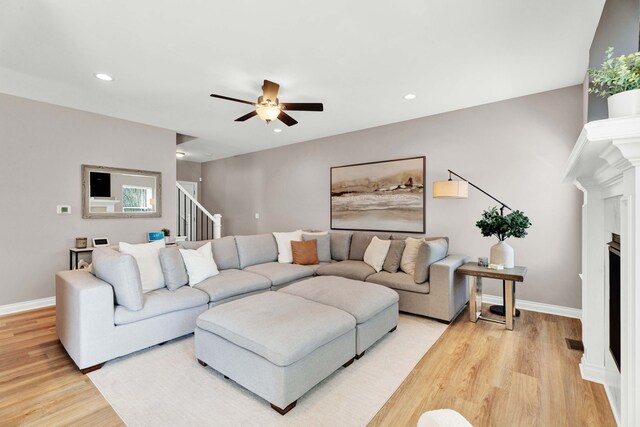 living area featuring stairs, a fireplace, ceiling fan, light wood-style flooring, and baseboards