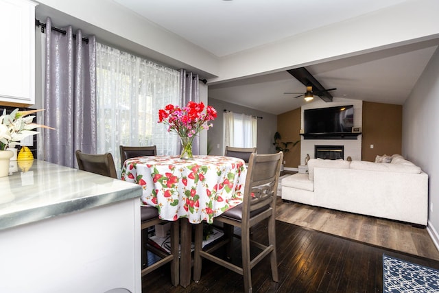 dining room featuring ceiling fan, a brick fireplace, baseboards, dark wood-style floors, and vaulted ceiling with beams