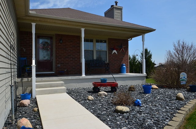entrance to property featuring a chimney, brick siding, a porch, and a shingled roof