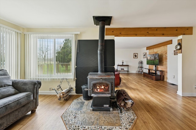 living area featuring baseboards, a wood stove, wood finished floors, and beam ceiling