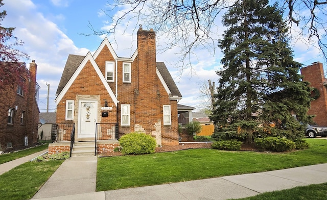 tudor-style house featuring brick siding, roof with shingles, a front yard, and a chimney