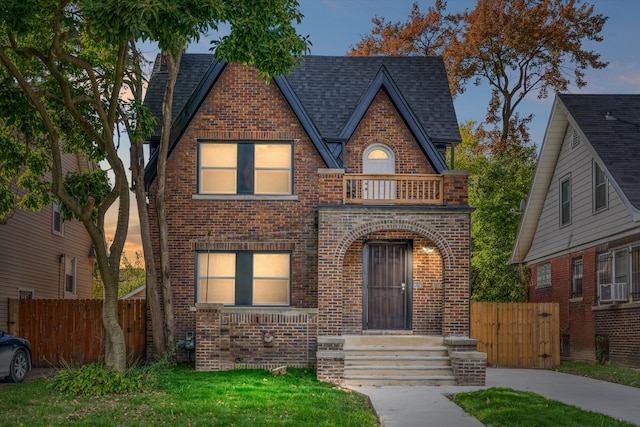 english style home with cooling unit, a balcony, a shingled roof, fence, and brick siding
