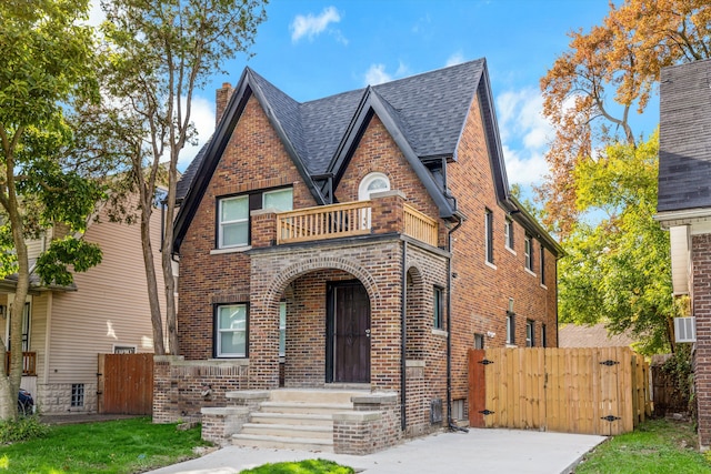 tudor-style house featuring a balcony, a gate, roof with shingles, fence, and brick siding