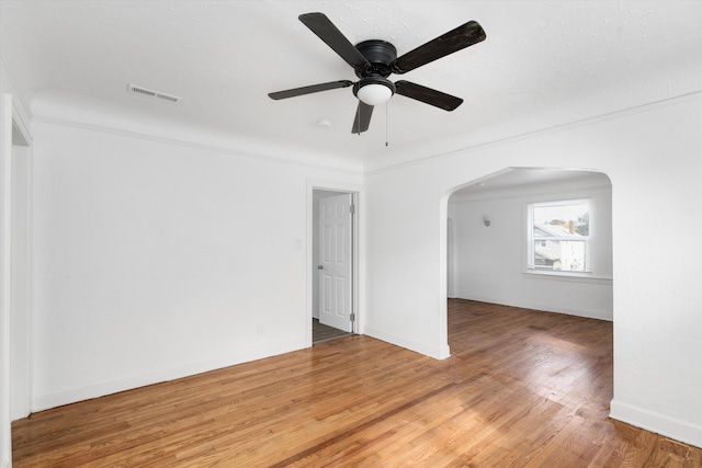 empty room with light wood-type flooring, a ceiling fan, visible vents, baseboards, and arched walkways