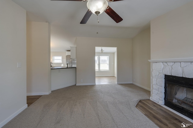unfurnished living room featuring carpet, a ceiling fan, a stone fireplace, and baseboards