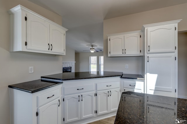 kitchen with dark stone counters, a ceiling fan, a stone fireplace, a peninsula, and white cabinets