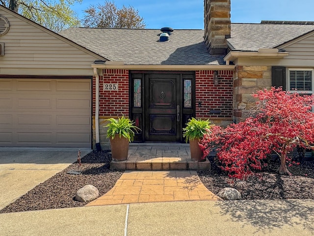 view of exterior entry with brick siding, roof with shingles, a chimney, and concrete driveway