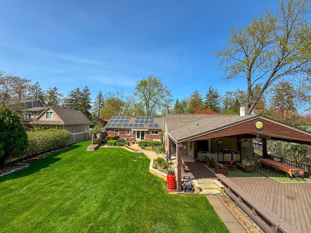 rear view of property featuring a shingled roof, a deck, roof mounted solar panels, a fenced backyard, and a yard