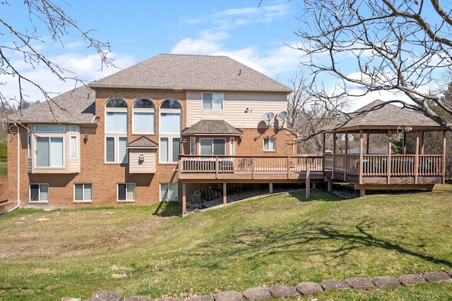 rear view of property with a gazebo, a deck, roof with shingles, a lawn, and brick siding