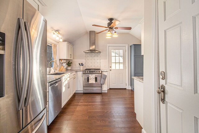 kitchen featuring a ceiling fan, a sink, light stone counters, stainless steel appliances, and wall chimney exhaust hood