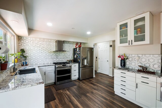 kitchen with wall chimney range hood, light stone counters, a sink, stainless steel appliances, and decorative backsplash