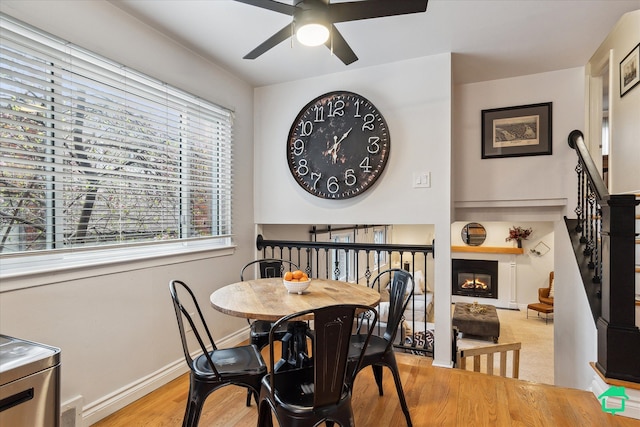 dining room featuring wood finished floors, stairs, ceiling fan, a glass covered fireplace, and baseboards