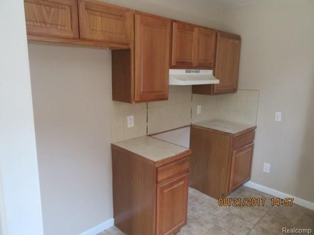 kitchen with tile counters, under cabinet range hood, brown cabinets, backsplash, and baseboards