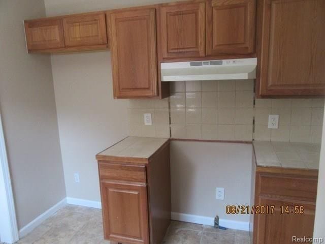 kitchen featuring brown cabinetry, tile counters, under cabinet range hood, and decorative backsplash