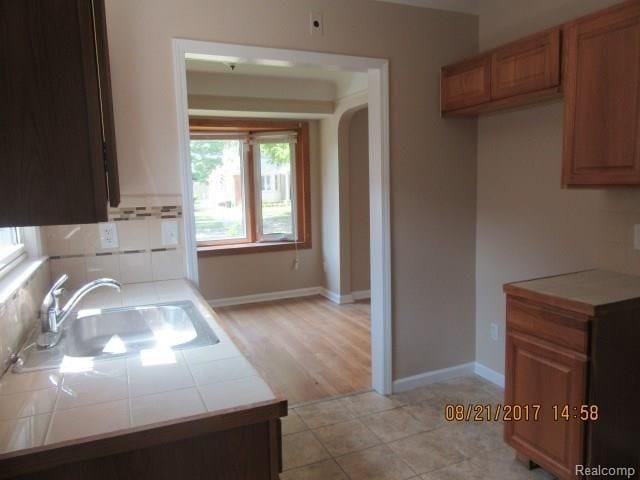 kitchen featuring arched walkways, decorative backsplash, brown cabinetry, a sink, and tile countertops