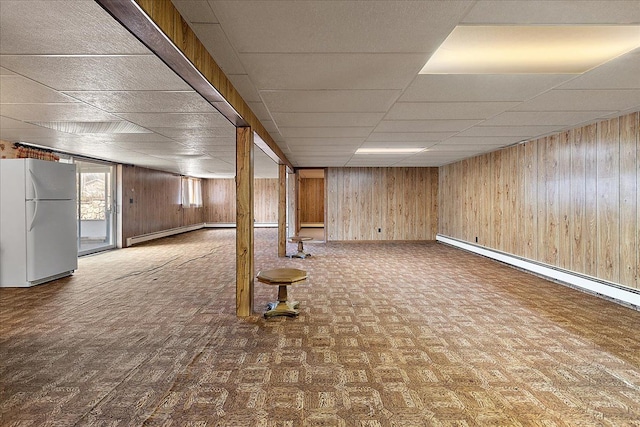 below grade area with baseboards, freestanding refrigerator, a drop ceiling, and wood walls