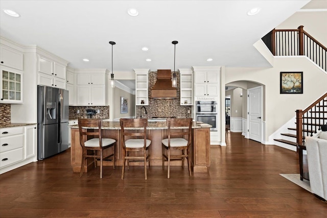 kitchen featuring light stone counters, glass insert cabinets, custom range hood, arched walkways, and stainless steel appliances