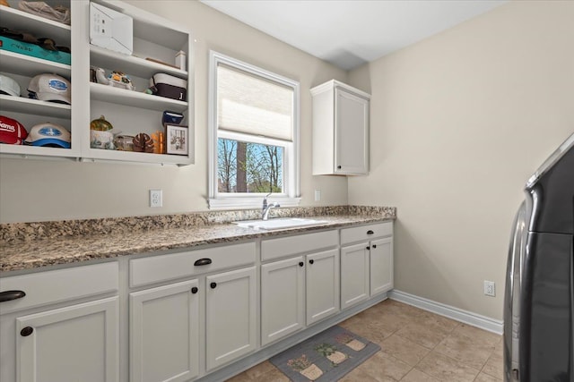 kitchen featuring refrigerator, a sink, baseboards, light stone counters, and white cabinetry