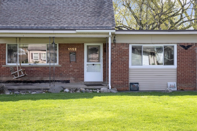 view of exterior entry featuring brick siding, a shingled roof, and a yard