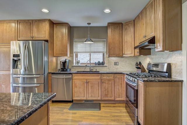 kitchen featuring appliances with stainless steel finishes, a sink, light wood finished floors, dark stone countertops, and under cabinet range hood