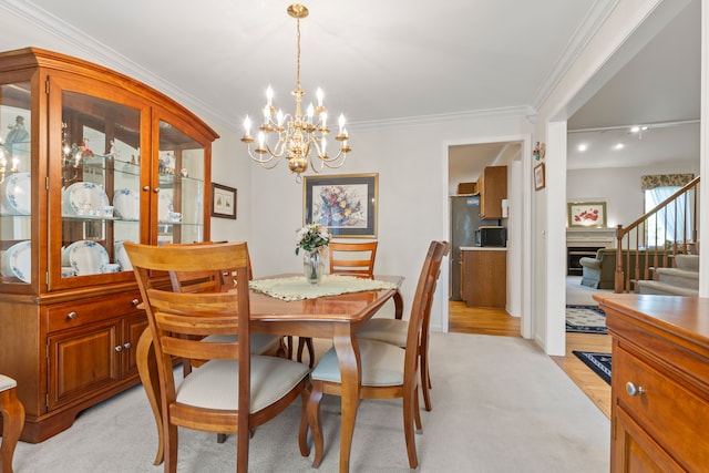 dining room featuring an inviting chandelier, ornamental molding, a fireplace, stairway, and light colored carpet