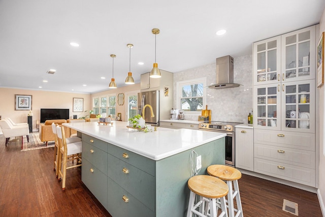 kitchen with wall chimney range hood, dark wood-style flooring, appliances with stainless steel finishes, an island with sink, and a kitchen bar