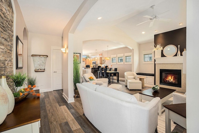 living area with a tiled fireplace, ceiling fan, vaulted ceiling, arched walkways, and wood finished floors