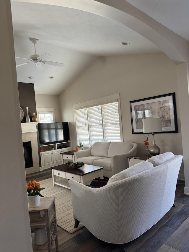 living room with a tiled fireplace, a ceiling fan, vaulted ceiling, arched walkways, and wood finished floors