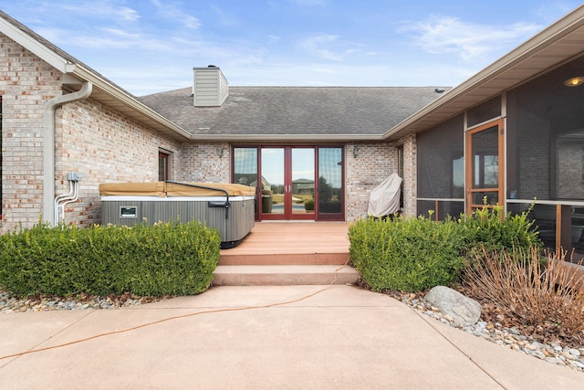 view of exterior entry featuring a chimney, brick siding, a shingled roof, a hot tub, and central AC unit