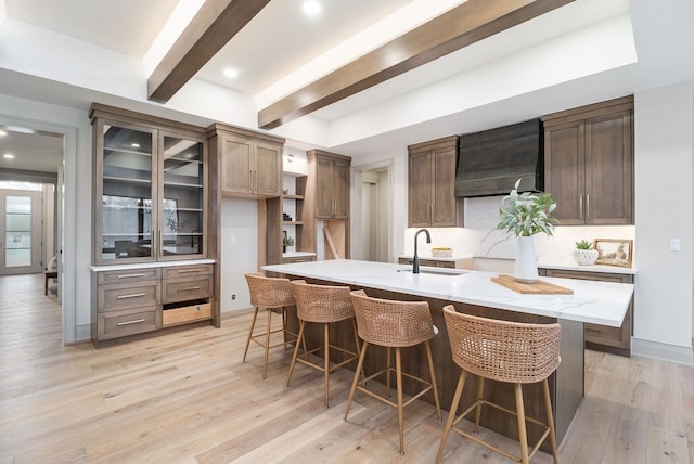 kitchen featuring custom exhaust hood, light stone countertops, glass insert cabinets, beamed ceiling, and a sink