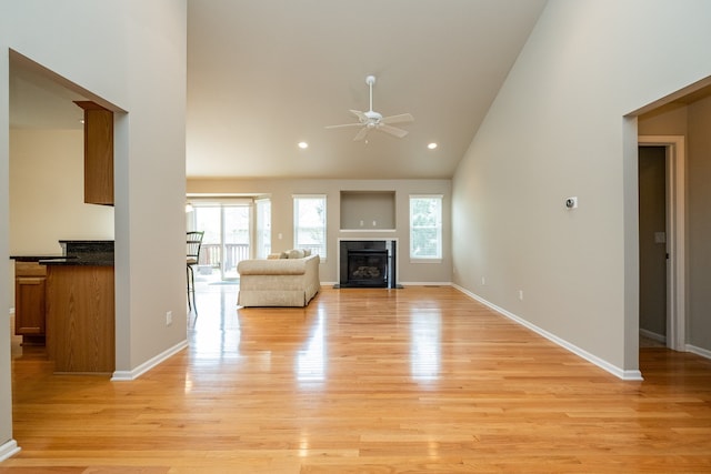 unfurnished living room featuring baseboards, ceiling fan, high vaulted ceiling, light wood-type flooring, and a fireplace with flush hearth