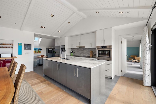kitchen featuring a center island with sink, lofted ceiling with beams, light wood finished floors, appliances with stainless steel finishes, and backsplash