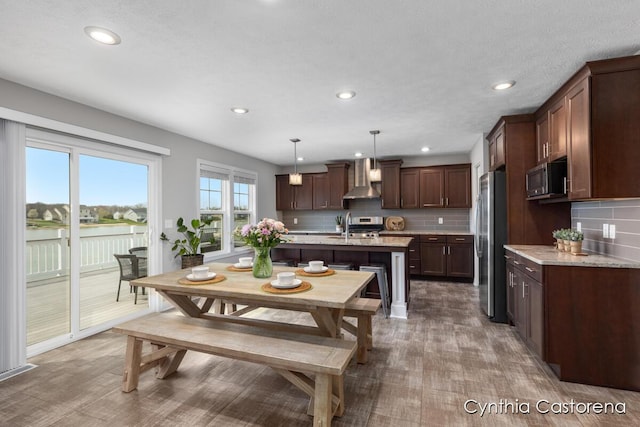 kitchen with stainless steel appliances, a center island with sink, wall chimney exhaust hood, and tasteful backsplash