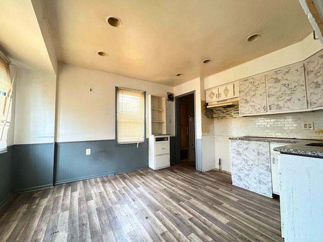 kitchen with dark wood finished floors, under cabinet range hood, backsplash, white cabinetry, and wainscoting
