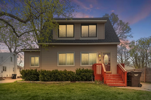 back of house at dusk with a shingled roof, fence, and a yard