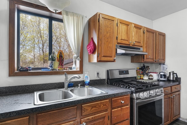 kitchen featuring brown cabinets, dark countertops, a sink, stainless steel gas range oven, and under cabinet range hood