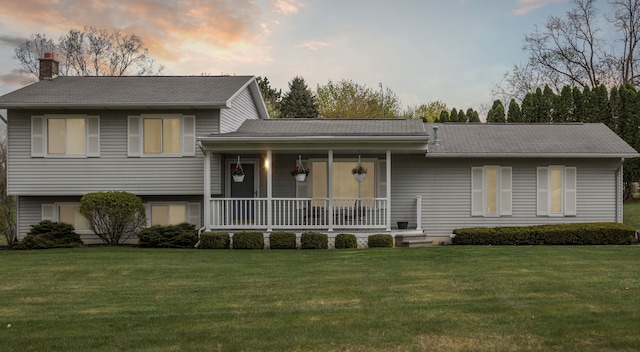 tri-level home featuring a shingled roof, covered porch, a chimney, and a yard