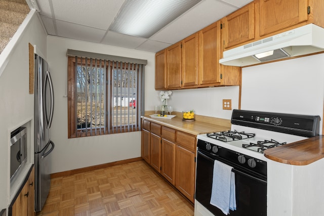 kitchen with brown cabinetry, a drop ceiling, light countertops, under cabinet range hood, and stainless steel appliances