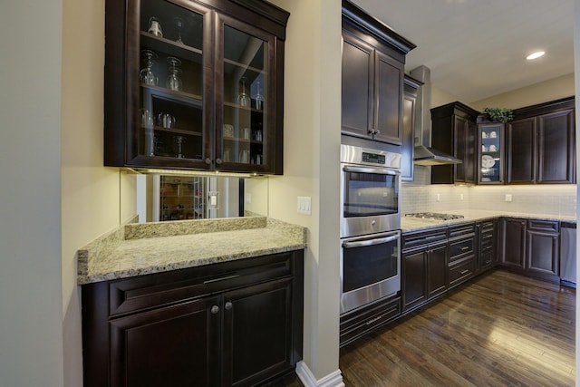 kitchen with wall chimney range hood, backsplash, dark wood-style flooring, light stone counters, and stainless steel appliances